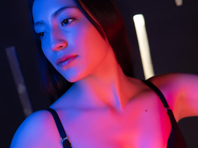 AnnFisher with blue and pink neon lighting, dark straight hair, black strap dress, soft expression, slight blush, small mole on right side of neck, collarbone visible.