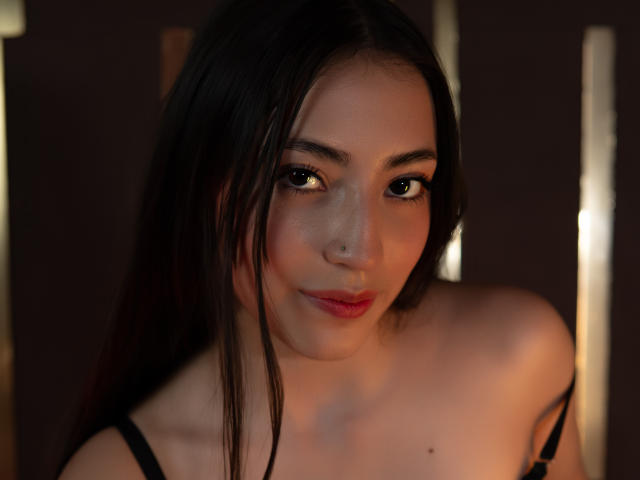 AnnFisher has long dark brown hair, a nose ring, and a subtle smile with pink lipstick. She's wearing a black strap top, showing her bare shoulders and tits.