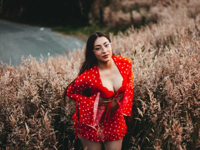 AnnFisher with long brown hair, wearing a red polka dot dress with a deep V, exposing her tits, standing in a field of tall, dry grass.