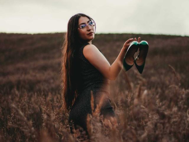 AnnFisher with long brown hair, wearing glasses and a black dress, holding green high heels in a brown field, looking over her shoulder with a slight smile.