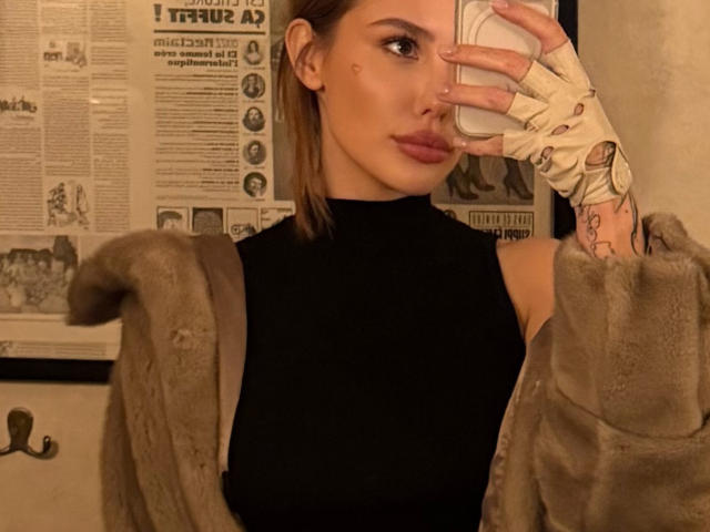 SensualMila, avec ses lèvres pulpeuses, portant un haut noir sans manches, un manteau en fourrure et un gant blanc à motifs, se prend en selfie, mettant en valeur ses tatouages et ses cheveux bruns..
