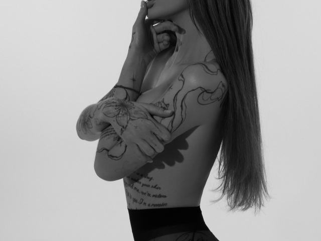 SensualMila con tatuajes en los brazos y costado, cabello largo y lacio, sin top, ropa interior negra, dedos cerca de la boca, foto en blanco y negro, brazos cruzados, mostrando pechos pequeños y un poco de busto..