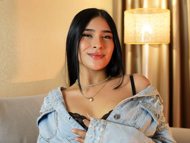 Antonella Preschot, avec ses cheveux noirs, sa peau claire et sa petite poitrine, porte une veste en jean portée asymétriquement, un soutien-gorge en dentelle noire et un collier de perles, et sourit doucement, debout devant une lampe..