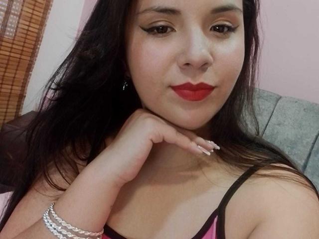 RosieTurner with long black hair, red lipstick, and black eyeliner, touching her chin with her hand, wearing a black and pink top, and a silver bracelet.