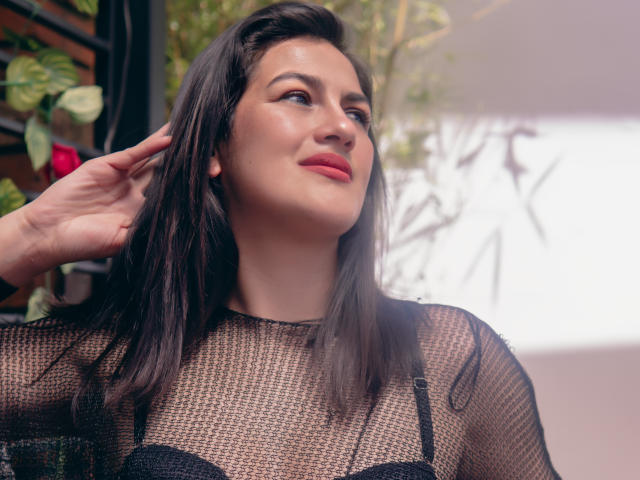 Elizabeth Dupont, con i capelli castano scuro, indossa una maglia nera trasparente che lascia intravedere il suo reggiseno nero, guarda verso l'alto con un leggero sorriso, con una mano che le accarezza i capelli..