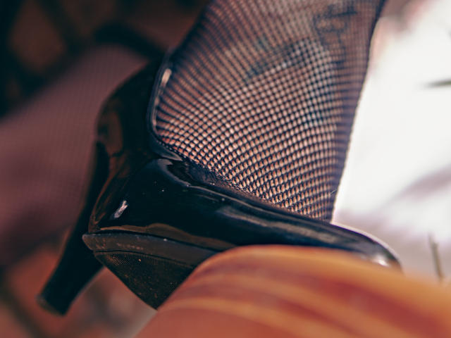 ElizabethDupont's shiny black stiletto with a fishnet top, showing a glossy heel and a wooden floor in the foreground. Tight, sexy, and classy.