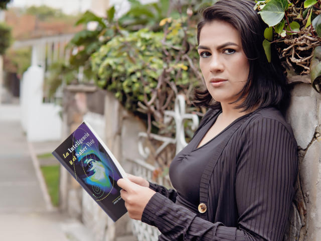 ElizabethDupont with short black hair, wearing a black cardigan and gray top, holding a book, standing against a wall with greenery, looking at the camera with a serious expression.