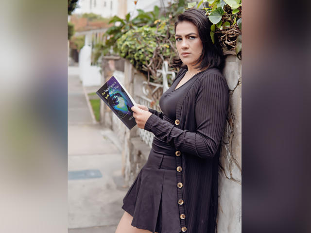 Elizabeth Dupont está de pie frente a una pared, sosteniendo un libro, vestida con un vestido negro con botones dorados y un cárdigan de punto acanalado, con su trasero ligeramente visible, cabello oscuro y una expresión seria..
