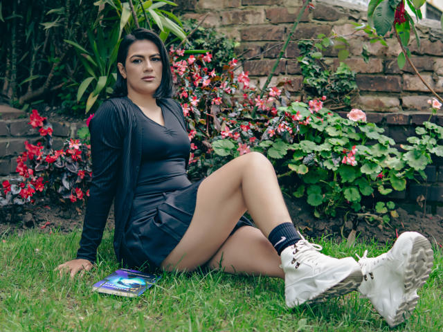 Elizabeth Dupont, met donker haar, een zwarte jurk en witte sneakers, zittend op het gras, benen gekruist, een boek op de grond, een bakstenen muur op de achtergrond, rode bloemen en groene planten in de achtergrond..