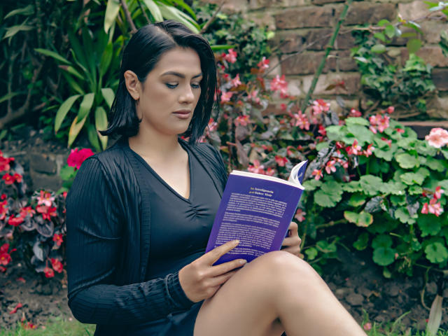 ElizabethDupont with dark hair, black cardigan, and black dress, reading a purple book, sitting with knees up, showing her ass, in a garden with red flowers.