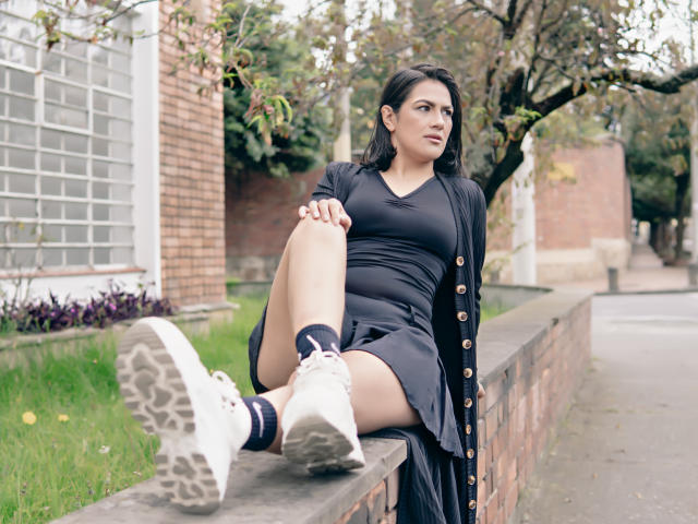 ElizabethDupont with dark hair, wearing a black dress and open cardigan, sitting on a brick wall, leg raised, white sneakers, black socks, looking to the side, outdoors.