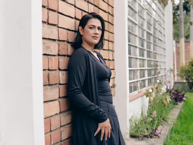 ElizabethDupont with black hair, medium tits, and a curvy ass, leaning against a brick wall in a black outfit, looking confident with a slight smile, right hand on her hip.