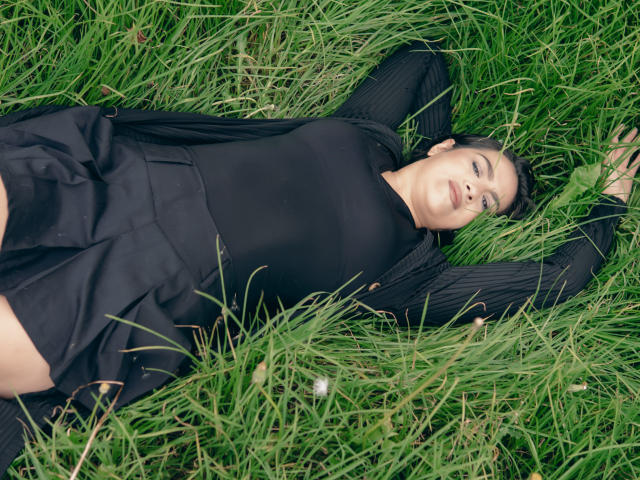 ElizabethDupont lies in green grass, arms above head, wearing a black top and skirt, with a relaxed, slightly smiling face, short dark hair, and a fair skin tone.