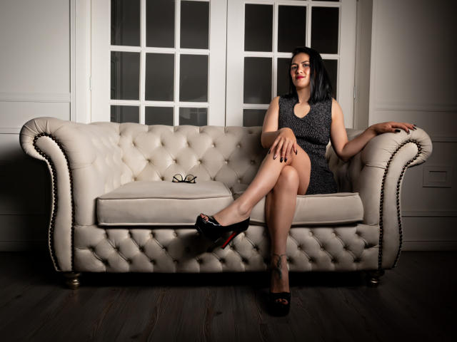 ElizabethDupont, with black hair, sits on a tufted white sofa, legs crossed, wearing a grey dress and black high heels, with red lipstick and a tattoo on her ankle.
