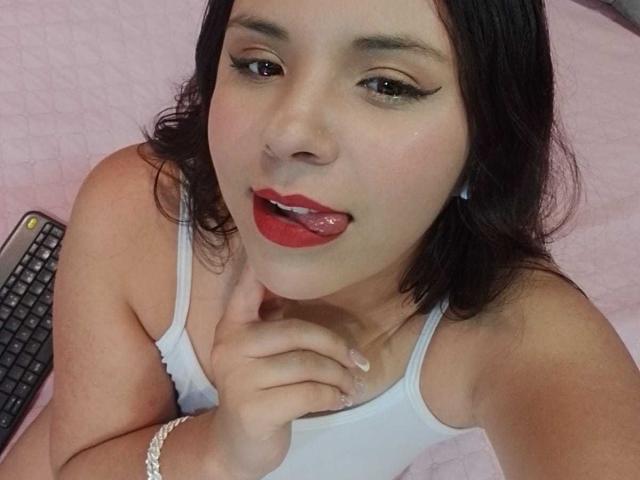 RosieTurner with red lipstick, black eyeliner, and tongue out, wearing a white tank top, finger near mouth, black keyboard in background, pink bed. RosieTurner has medium-length black hair.