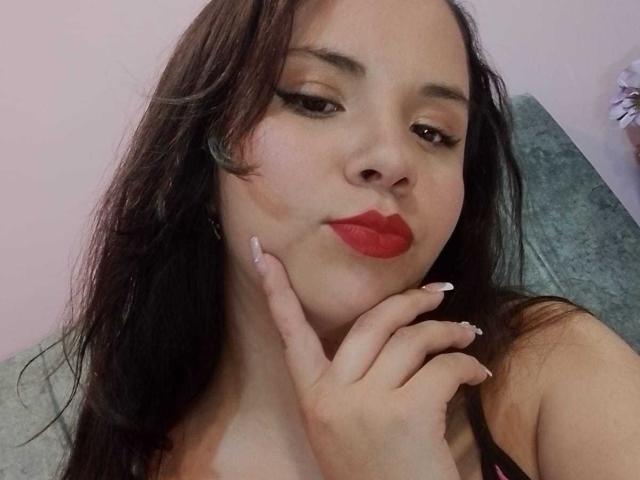 Close-up of RosieTurner. She has fair skin, long dark brown hair, and red lipstick. Her right hand, with white, long, pointed nails, touches her chin. She has subtle, winged eyeliner. The image focuses on her face and upper chest, showing a slight, confident smile. She wears a dark-colored top with thin straps. The background is blurred, with a light pink wall and a grey, textured surface. Her expression is relaxed and slightly seductive. The overall composition is intimate and detailed.