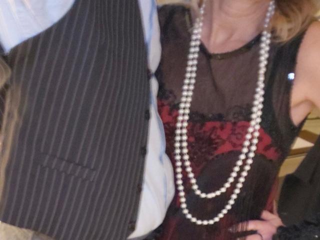 Close-up of HotSexyPaar-hot's upper body. Wearing a pinstriped black vest over a light blue shirt, with a sheer black top that has red lace detailing. Long pearl necklace hangs down to their chest. HotSexyPaar-hot's right arm is visible, with their hand resting on their hip. The image is slightly blurred, emphasizing the textures of the fabric and the sheen of the pearls. The color contrast between the black, red, and white is prominent.