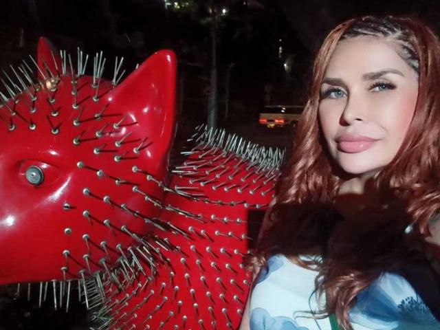 Xuxaanall with red, spiked fish sculpture, long wavy brown hair, light skin, and green eyes, wearing a blue and white top, smiling at the camera.