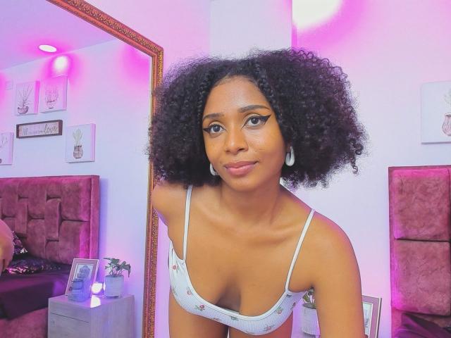 MeganWlliams with curly black hair, wearing a white, floral-patterned, spaghetti-strap top, showing her small, brown, perky tits, standing in front of a mirror, smiling softly.