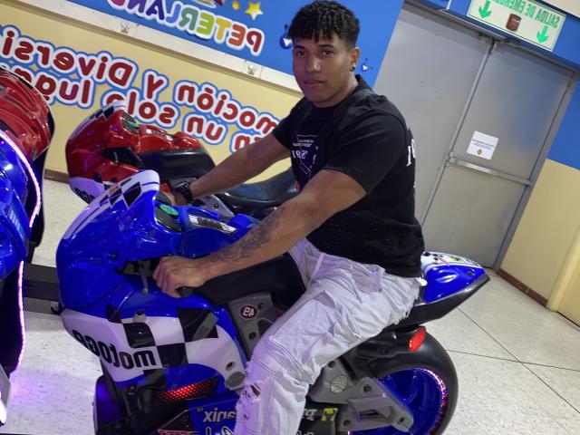 Yoximanuelprime, con cabello negro rizado y corto, vistiendo una camisa negra y pantalones blancos, está sentado en una motocicleta azul Yamaha, agarrando el manillar con una expresión seria..