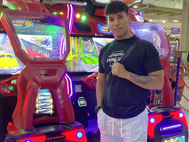 Yoximanuelprime está de pie frente a máquinas de arcade de colores, vistiendo una camiseta negra y pantalones cortos blancos, con un tatuaje en su brazo derecho y una ligera sonrisa..