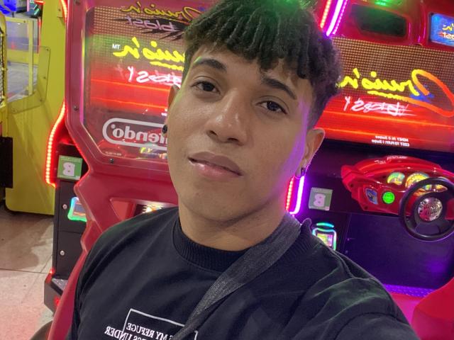 Close-up selfie of Yoximanuelprime. Yoximanuelprime is a young man with medium brown skin, short curly black hair, and a slight smile. He wears a black hoodie with white text on the front. The background features a brightly lit, neon-red arcade machine with Arabic writing and colorful buttons. The machine has a steering wheel on the right and is illuminated by purple and yellow neon lights. The image has a slight green filter on the top left. Yoximanuelprime is centered, looking directly at the camera.