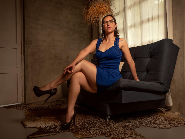 AndromedaJade in a blue dress, sitting on a black chair with her legs crossed, wearing black high heels, looking at the camera with glasses, and a serious expression.