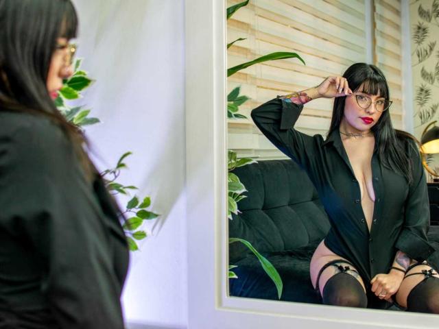 KuroBabee with black shirt, glasses, and red lipstick, sitting on black sofa, reflecting in mirror, showing garters and black stockings, with her hand near her head.