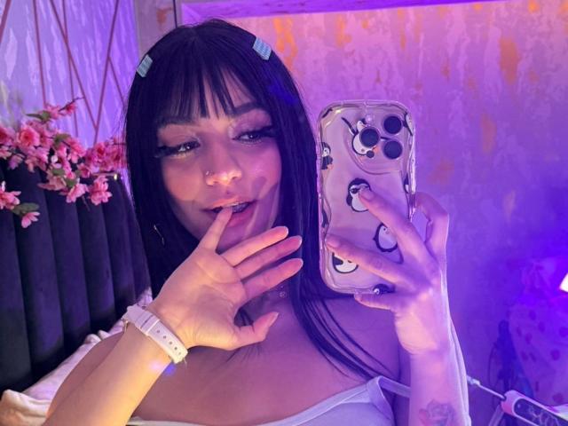 LindaWatsong with black hair, blue hair clips, and white tank top, holding phone, finger near mouth, purple and pink lighting, white wristband, floral decor, visible tattoo.