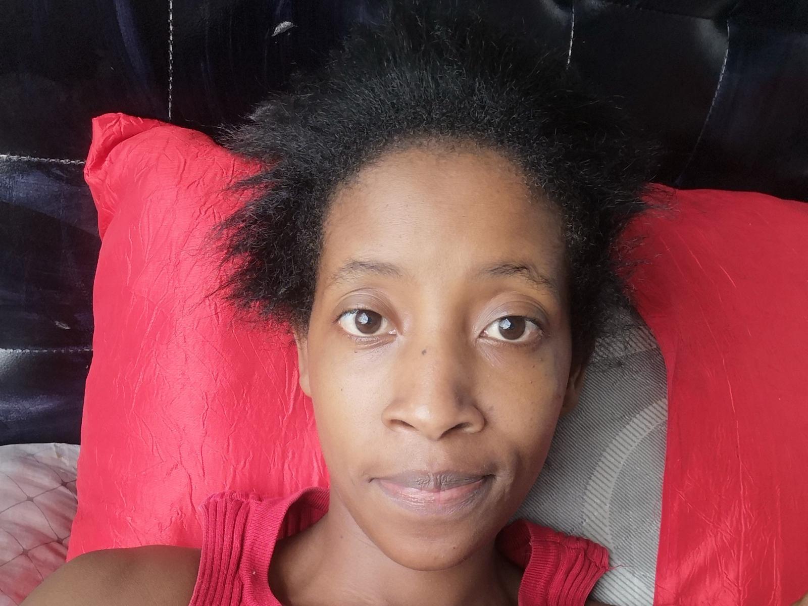 TrishJacquelyn's face is centered in the image. She has medium brown skin, brown eyes, and a small mole on her right cheek. Her black, curly hair is slightly messy. She's wearing a red sleeveless top. The background includes a red pillow on the left and a grey pillow on the right. The headrest behind her is dark and textured. The image has a realistic, natural style. Her expression is neutral, and she's looking slightly to the right.