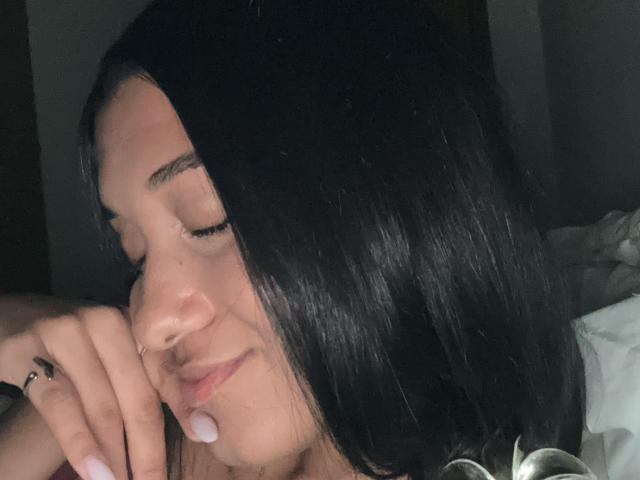 Close-up of MilenaZ with closed eyes and a slight smile. She has long, straight black hair that partially covers her face. A small, white drop of saliva is on her lower lip. Her right hand, with a black ring on her index finger, rests on her cheek. The lighting is low, highlighting her smooth skin and relaxed expression. The background is dark and blurry, keeping the focus on MilenaZ's face and hair. The image has a soft, natural texture.