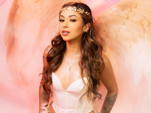 LilithMalkova stands against a soft pink backdrop with large, semi-transparent wings. She has long, wavy brown hair and wears a gold leaf headband. Her makeup is bold with dark eyeliner and pink lipstick. She's dressed in a white, strapless corset that emphasizes her cleavage. A gold heart necklace and matching earrings are visible. Her tattoos on both arms are detailed. The image has a warm, ethereal quality with subtle lighting. Her expression is confident and slightly seductive.