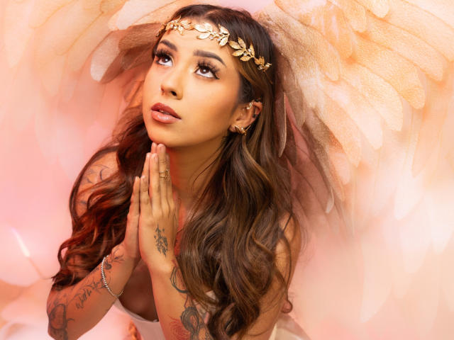 LilithMalkova, com cabelos castanhos longos, uma coroa de folhas douradas e asas de anjo, olhando para cima com as mãos perto do rosto, mostrando tatuagens na mão direita e no pulso..