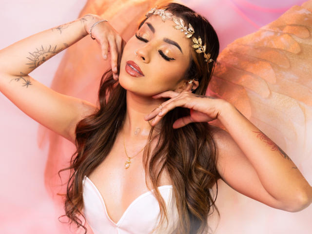 LilithMalkova with long, wavy brown hair wears a gold leaf headpiece and white strapless top. She has closed eyes, glossy pink lips, and a serene expression. Her hands touch her face, revealing tattoos on her left arm. She has a gold necklace with a small pendant. Angel wings, semi-translucent and orange, are visible behind her. The image has a soft, pinkish background. She has a light brown skin tone and is styled with a sensual, ethereal vibe.