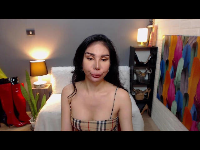 AsianTsGoddess with long black hair, plaid top, small tits, pouty lips, sitting in front of a lamp and colorful wall art, looking cute and slightly playful.