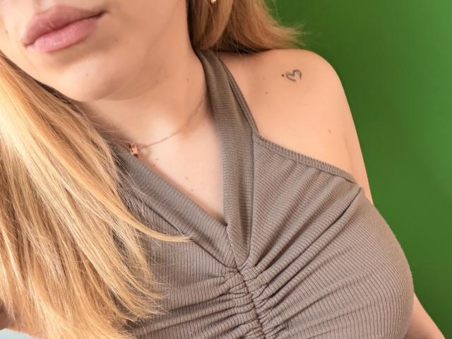 SugarrX with blonde hair, pink lips, and a small heart tattoo on the right shoulder, wearing a ribbed, gray halter top, showing her chest and neck.