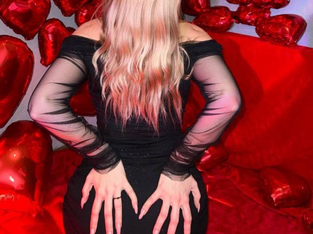 CrystalHope's back, hands on her ass, wearing a black off-shoulder dress with sheer sleeves, blonde hair, red heart balloons, red couch, in a sexy, romantic setting.