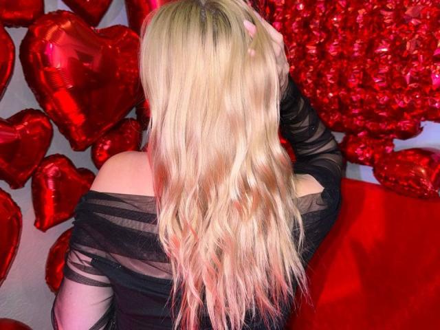 CrystalHope with long blonde hair, wearing a black off-shoulder top, has her hand in her hair, surrounded by red heart balloons. Her ass is partially visible.