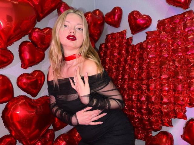 CrystalHope with blonde hair, wearing a sheer black off-shoulder dress, red choker, and red heart balloons, showing her tits and arms, in a sexy, red-themed setup.