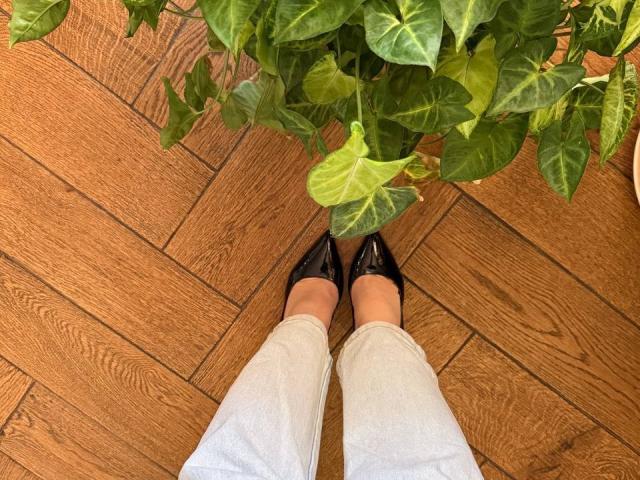 KaiaLein's foot in a shiny black stiletto with red soles, showing off a sleek heel against wooden floor and white jeans. Her other foot is in a black pointe.