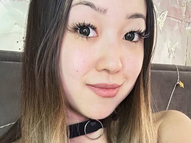 Close-up photo of RenyLime with fair skin and long, straight brown hair with blonde highlights. RenyLime has large, dark eyes with long, thick eyelashes and pink, slightly pursed lips. She wears a black collar with a silver ring around her neck. Her eyebrows are thin and slightly arched. The background features a muted, patterned wall with small, white butterfly designs. The image focuses on RenyLime's face, highlighting her subtle smile and relaxed expression. The photo is brightly lit, emphasizing her clear complexion and soft, natural makeup.