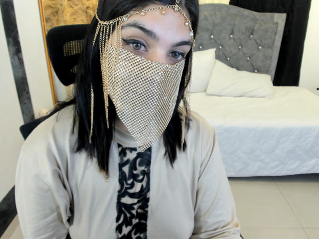 AniisaAli has straight black hair, wearing a gold chain headpiece and a gold mesh face mask with hanging chains. She has light skin and dark, defined eyebrows. Her eyes are blue. She wears a beige cardigan over a black and white patterned shirt. She's sitting in front of a bed with a tufted gray headboard and white pillows. The room has a wooden door and tiled floor. The image focuses on her upper body and face.