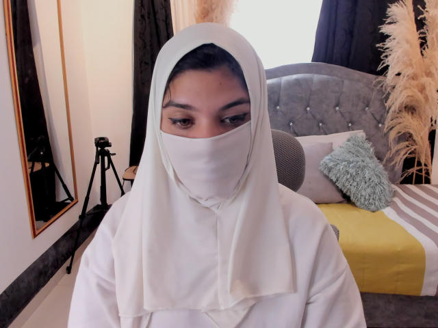 AniisaAli, wearing a white hijab and matching face cover, sits in a bedroom. She has medium brown skin and dark eyebrows. The background includes a gray tufted headboard, a yellow and gray bedspread, a fluffy green pillow, and pampas grass. A black tripod stands to the left, and a mirror is partially visible. The image focuses on AniisaAli's face and upper body, with a soft, natural light.