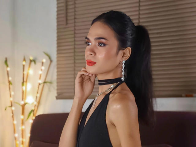 PeachAnderson-hot with black hair in a ponytail, wearing a black halter top, red lipstick, and a choker, standing with hand near chin, looking to the side.