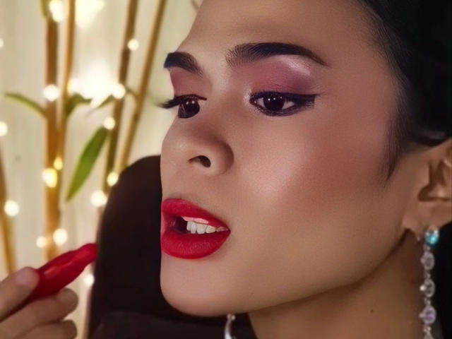 PeachAnderson-hot's face with dark eyebrows, purple eyeshadow, red lipstick, and glossy red lips being touched by a red lipstick. She has a light skin tone and black hair.