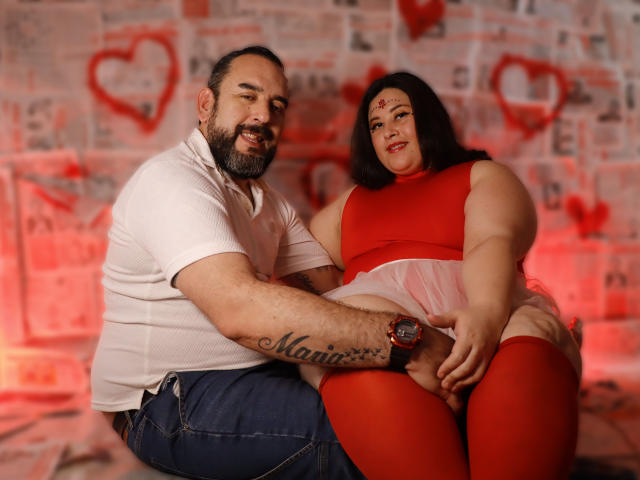 SarahMat, una donna robusta con un vestito rosso e calze alte, è seduta in grembo a un uomo. Lui ha la barba, indossa una camicia bianca e jeans blu, e ha un tatuaggio sul braccio. Sullo sfondo ci sono cuori rossi..