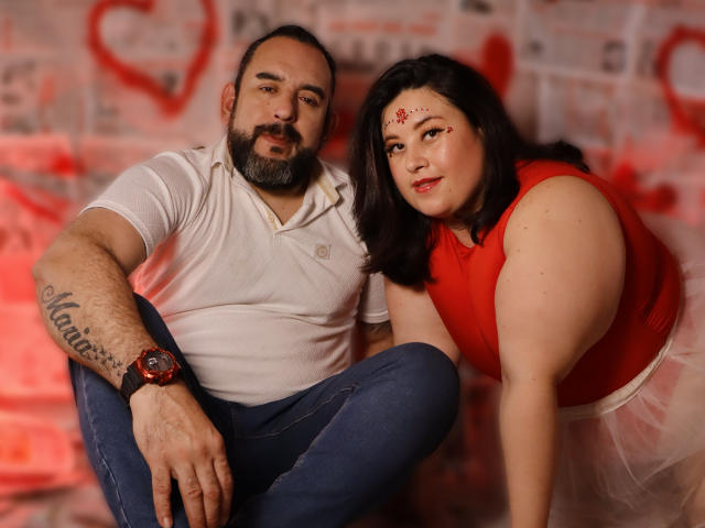 SarahMat met donker haar, een rode top en een witte rok, hurkt naast een man met baard in een witte polo, blauwe jeans en een rode horloge. Beide kijken naar de camera, en SarahMat heeft rode make-up..