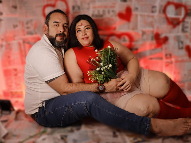 SarahMat, con barba, camisa blanca y jeans azules, está sentada junto a una mujer curvilínea con un vestido rojo, sosteniendo un ramo de flores, sobre un suelo cubierto de periódicos, con corazones rojos en el fondo..