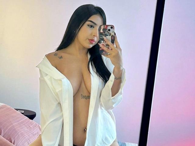 BellaVibes with long black hair, open white shirt revealing big tits, tattoos on shoulder and chest, holding phone, pink pillow in background, soft pink and purple lighting.