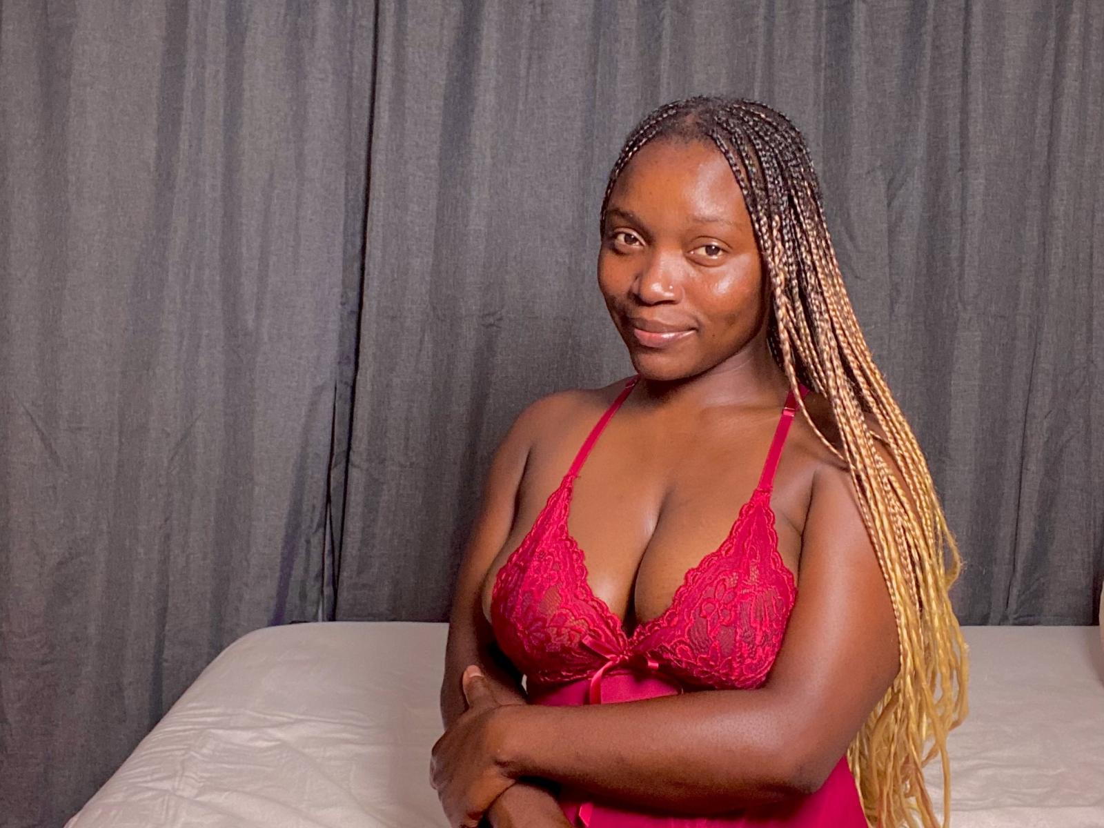 TaBelleDiwa with dark brown skin, long blonde braids, red lace lingerie, showing her boobs, arms crossed, smiling at the camera, standing in front of a gray curtain.